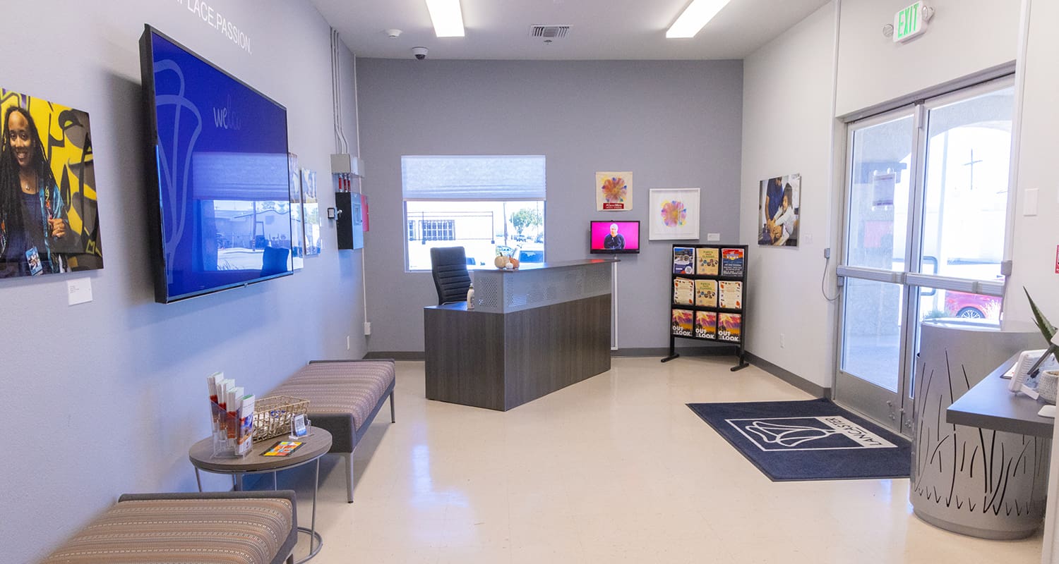 A waiting area features a reception desk, a TV, two chairs with a small table, magazines, and wall art. A window and glass door provide natural light.