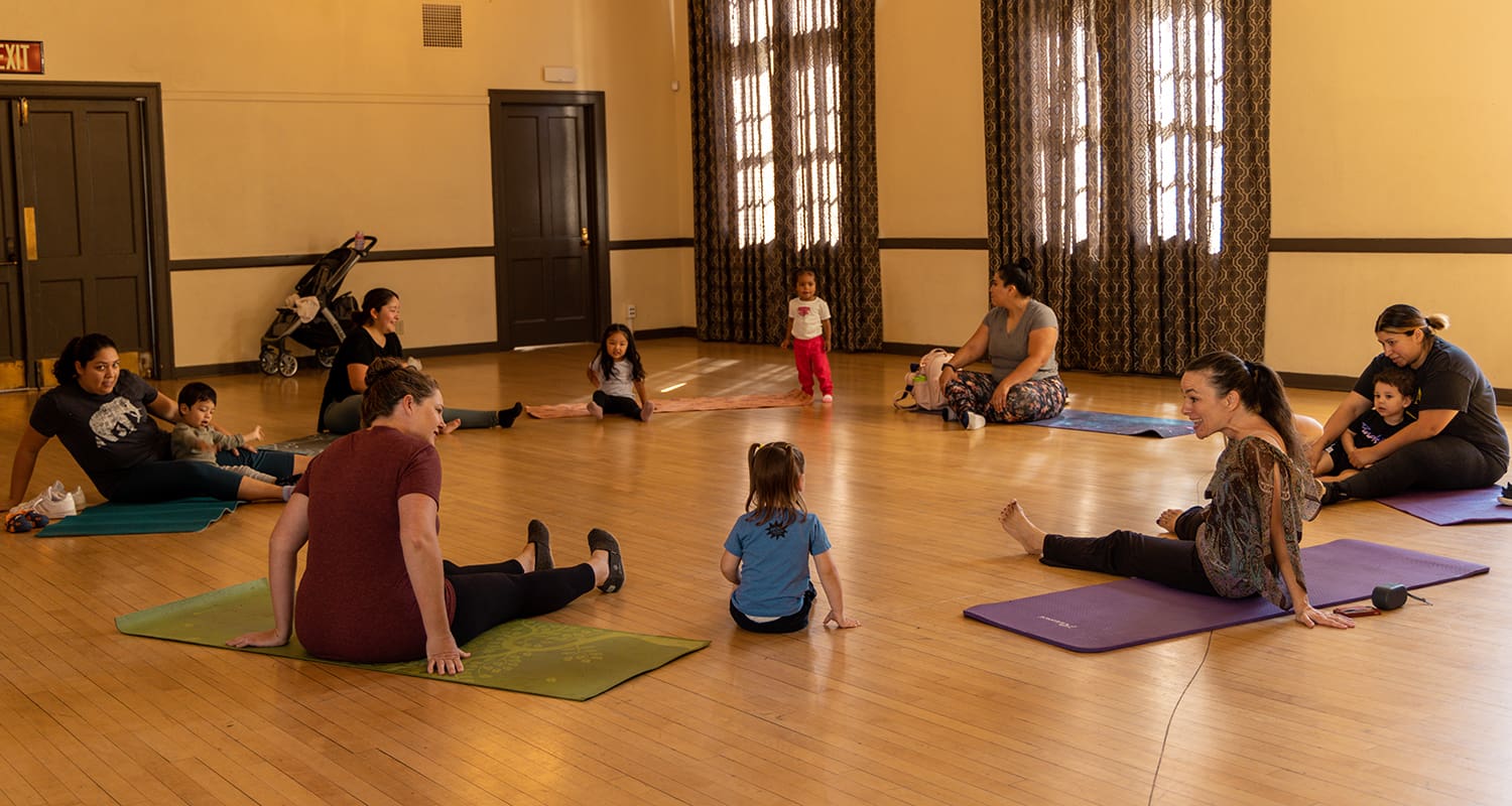 Adults and children sit on yoga mats in a circle in a room with wooden floors and large windows.