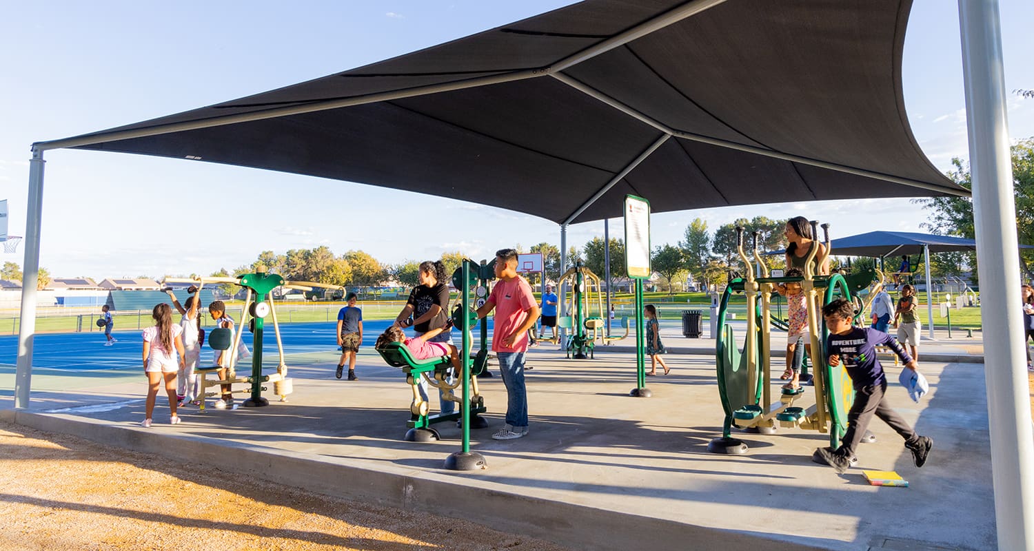 People are utilizing outdoor fitness equipment under a large shade structure in a park. Some children are playing nearby on a blue surface area, while others are exercising with the equipment.