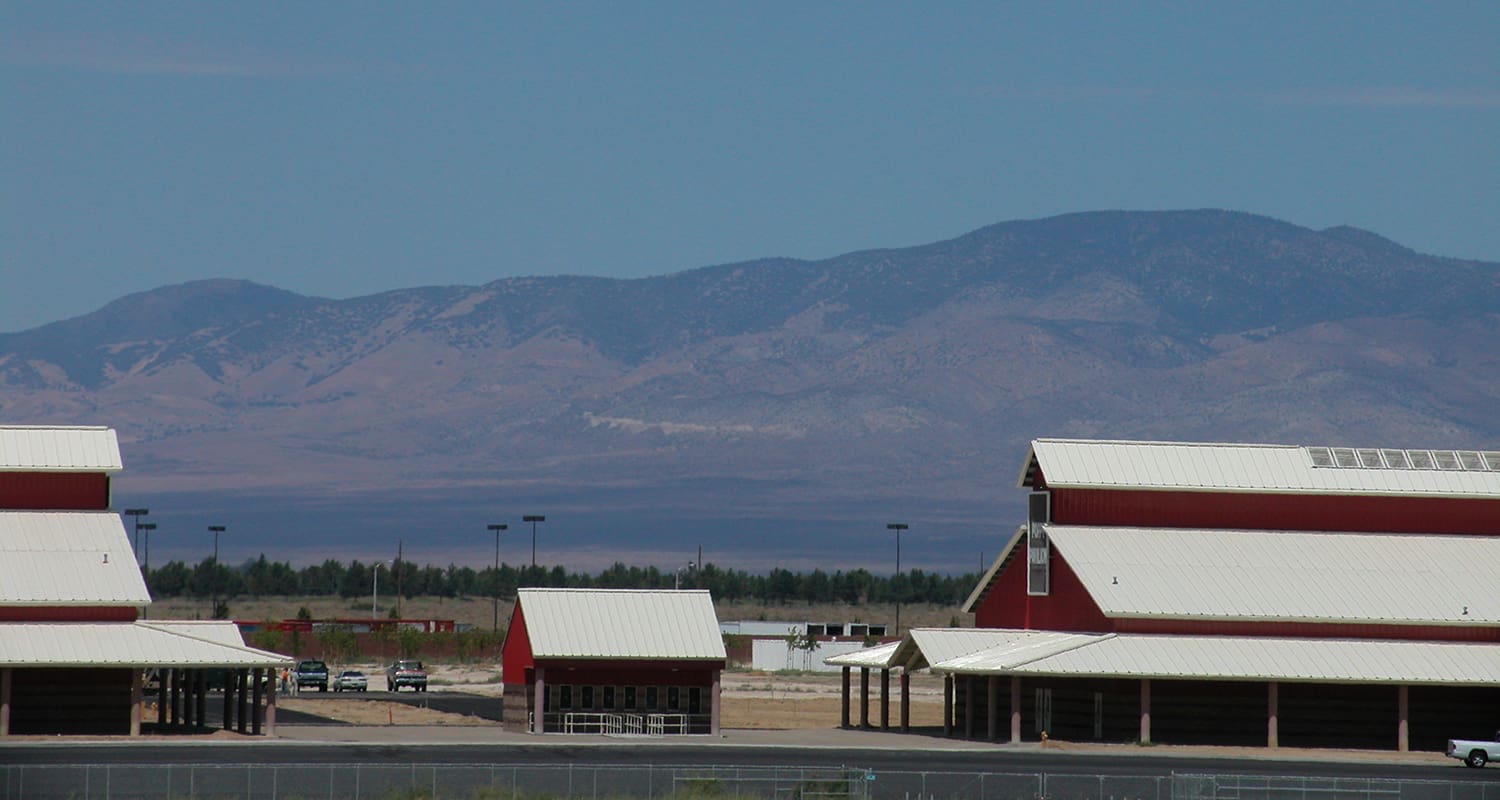 Red barn-like buildings with white roofs are in the foreground, while a mountain range under a clear sky stretches across the background. Some vehicles are visible behind a fence near the buildings.