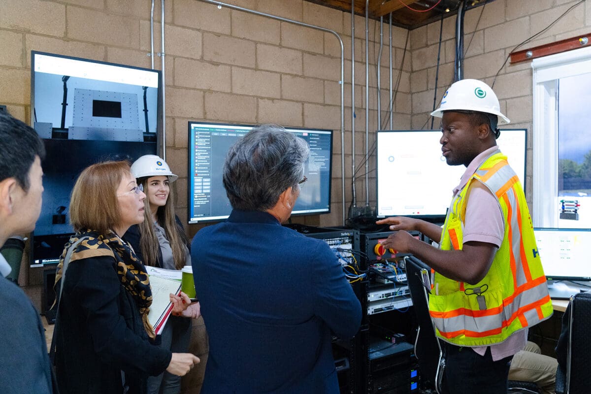 A person in a high-visibility vest and hard hat speaks to a group of people in an office setting with multiple monitors and a brick wall in the background.