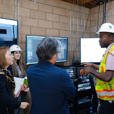 A person in a high-visibility vest and hard hat speaks to a group of people in an office setting with multiple monitors and a brick wall in the background.