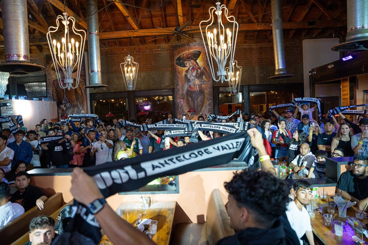 A crowd of fans inside a lively indoor venue hold up black and white scarves reading "1955 ANTELOPE VALLEY." Loft-style ceiling, large lights, and a mural featuring a cowboy in the background.