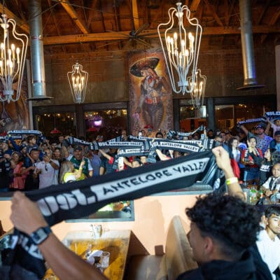 A crowd of fans inside a lively indoor venue hold up black and white scarves reading "1955 ANTELOPE VALLEY." Loft-style ceiling, large lights, and a mural featuring a cowboy in the background.