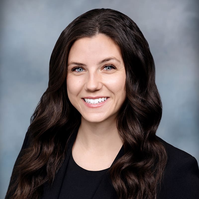 Portrait of a woman with long wavy brown hair, wearing a black blazer, smiling against a blurred gray background.