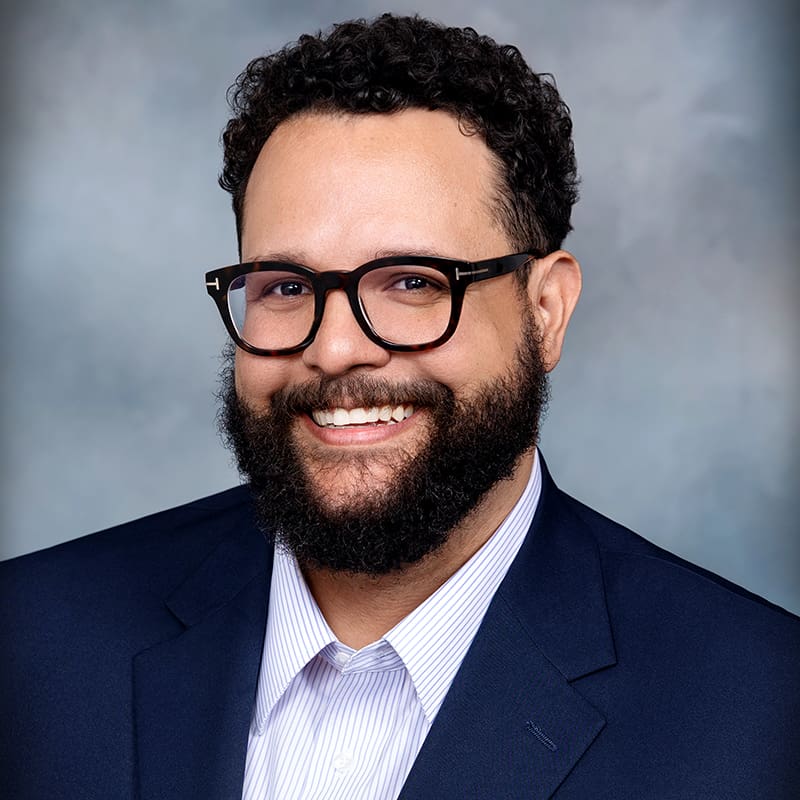 A bearded man with curly hair and black glasses is smiling, wearing a dark blue suit jacket over a light blue shirt, against a neutral background.