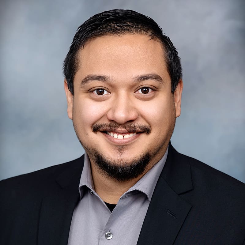 A man with short dark hair and a beard, dressed in a gray shirt and black blazer, smiles against a gray-blue background.