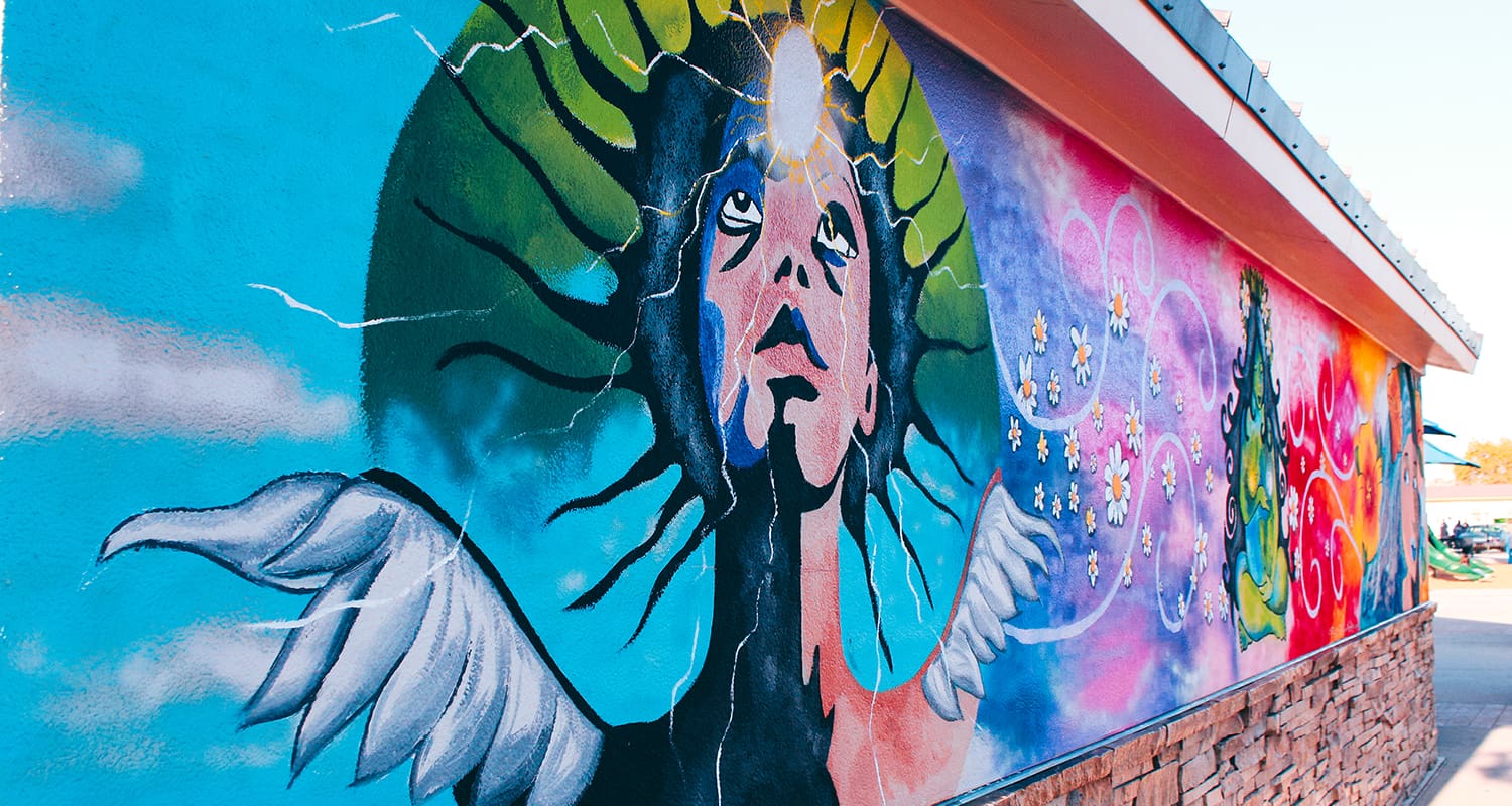 A colorful mural on a building wall features a person with wings looking upward, surrounded by vibrant, abstract elements on a bright day.