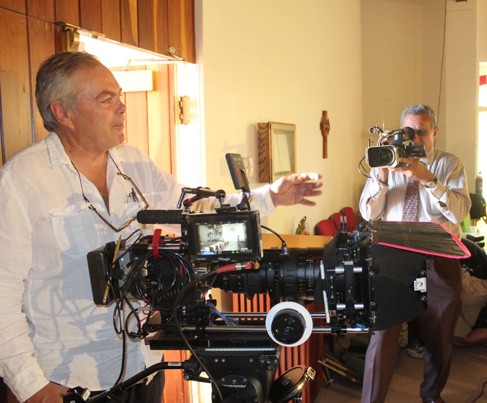 Two men are engaged in filming. One operates a professional camera setup, while the other captures the scene with a smaller video camera.