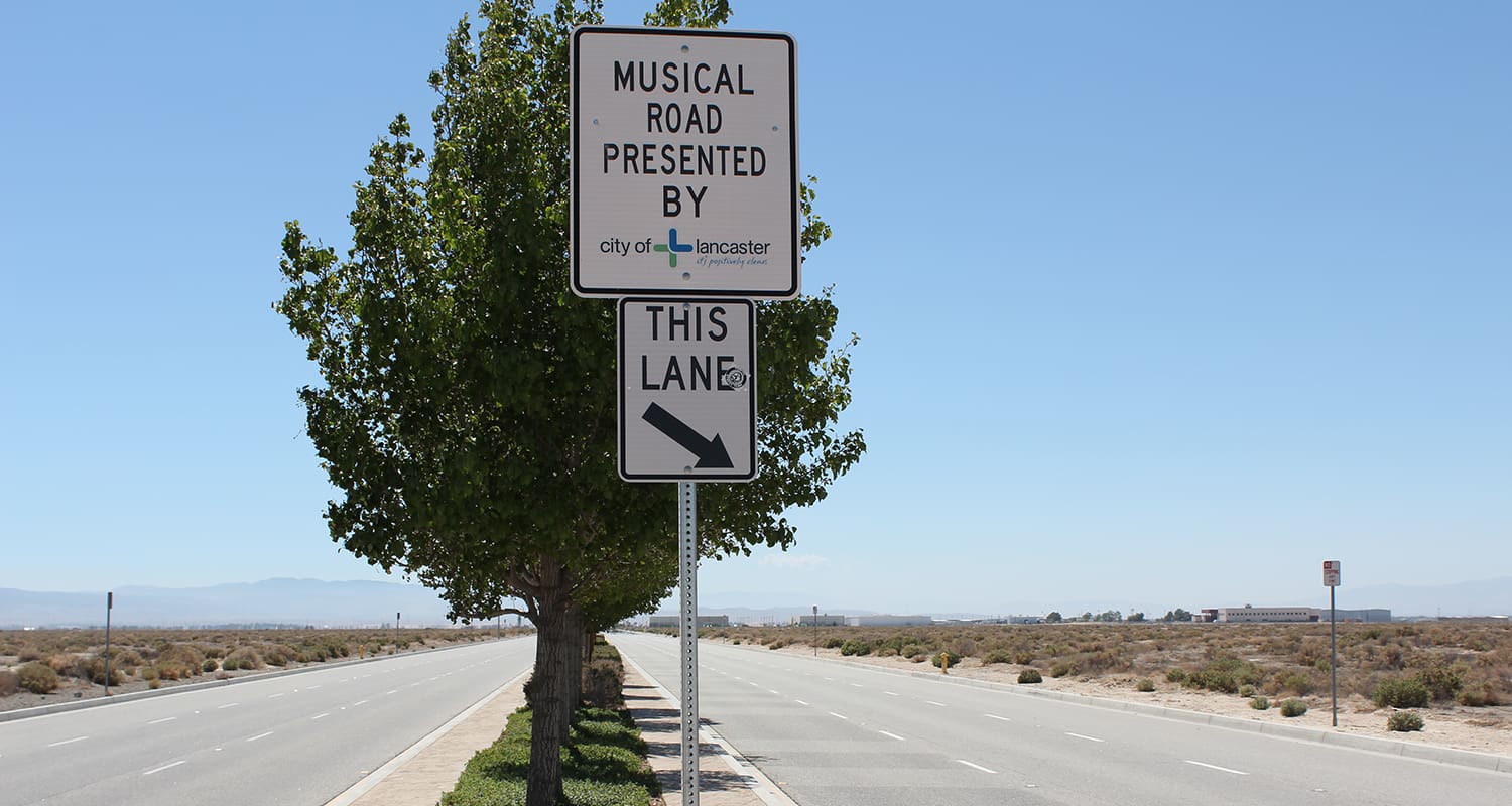 Road sign reading "Musical Road presented by City of Lancaster" with an arrow pointing right, next to a tree in a clear, open area.