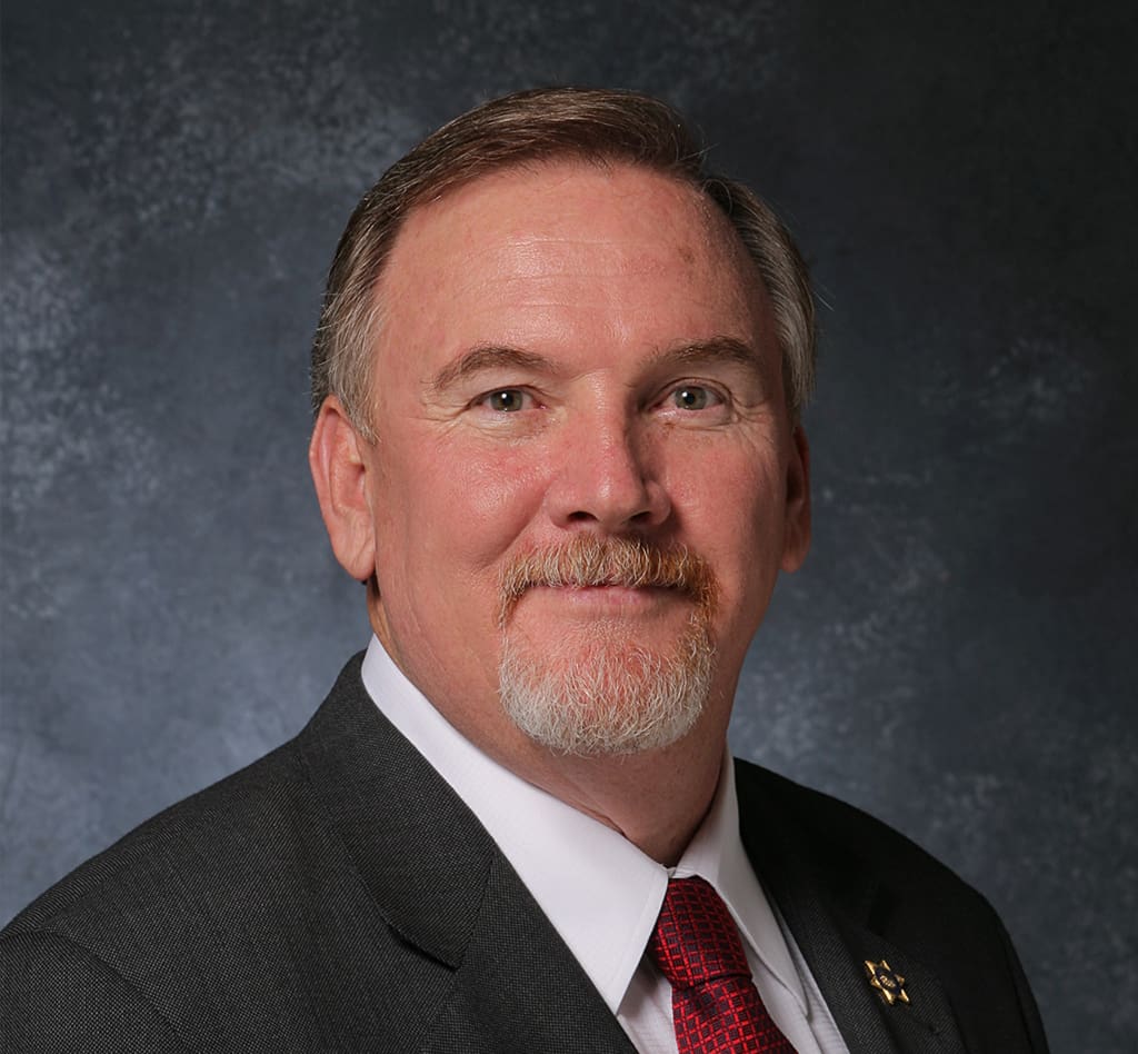 A man with short, light-colored hair and a white beard and mustache is wearing a dark suit, white shirt, and red tie, posing against a dark textured background.