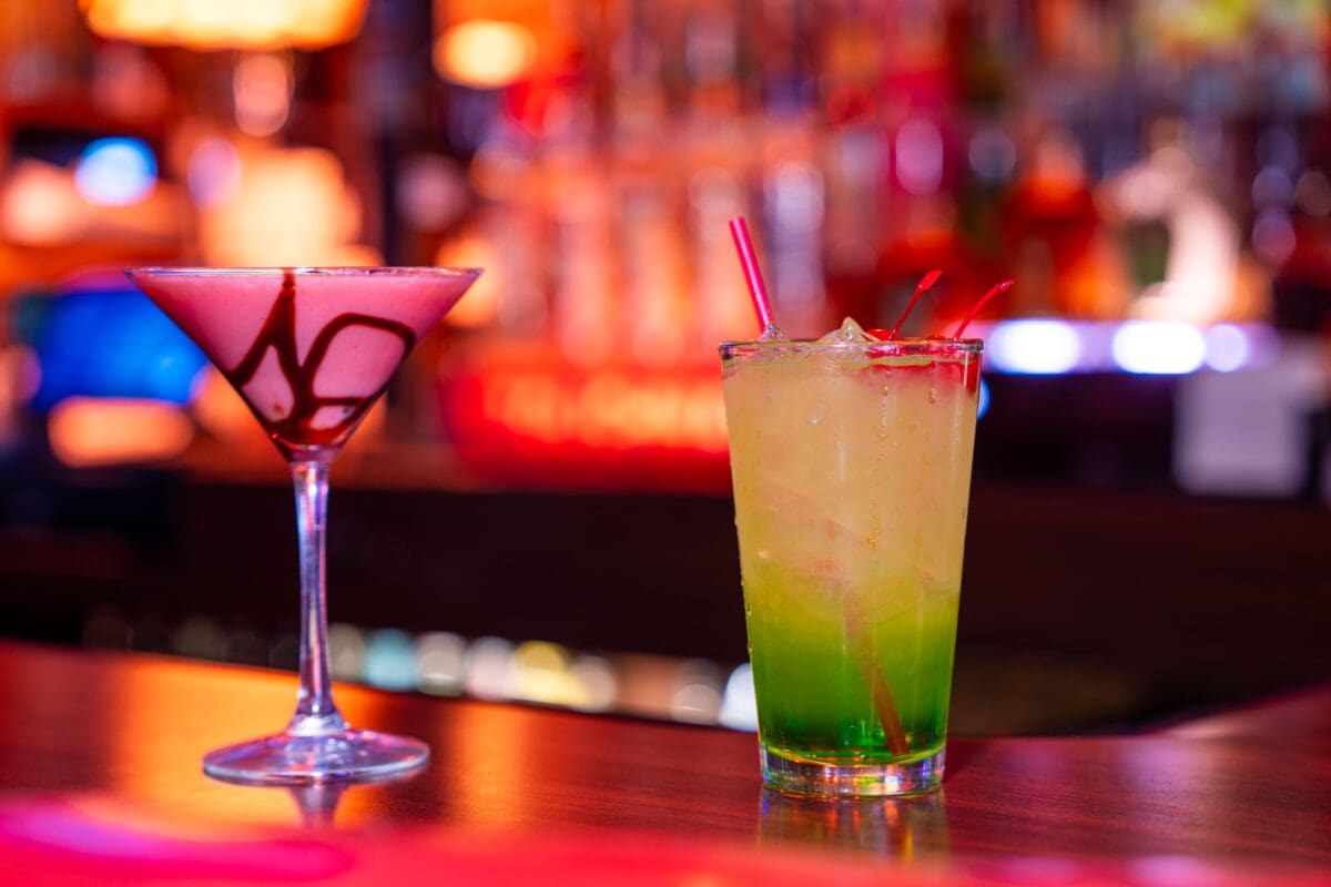 A martini glass with a chocolate dessert cocktail and a tall glass with a green cocktail and red straw on a bar counter.