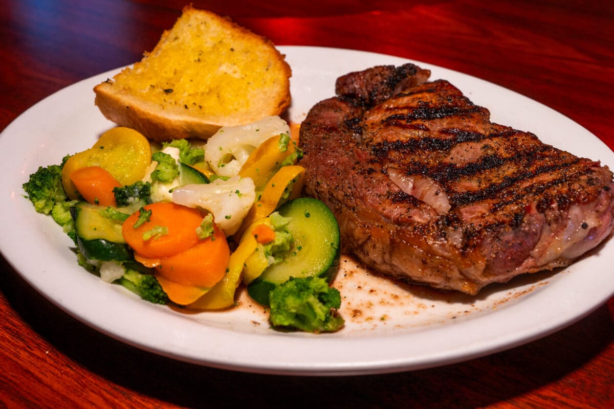 Grilled steak with grill marks on a white plate, accompanied by steamed mixed vegetables and a slice of toasted garlic bread on a wooden table.