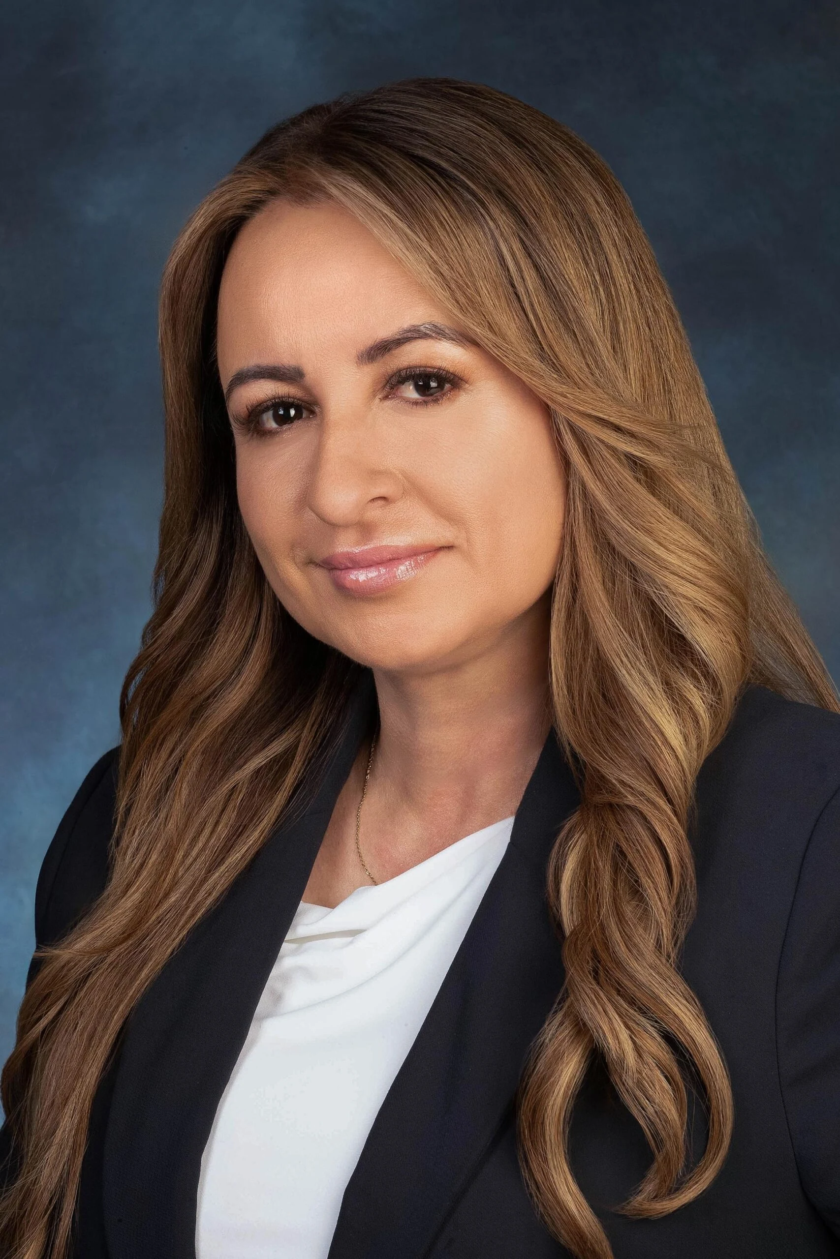 Professional headshot of a city councilmember with long light brown hair, wearing a black blazer over a white blouse, posed against a blue gray studio backdrop
