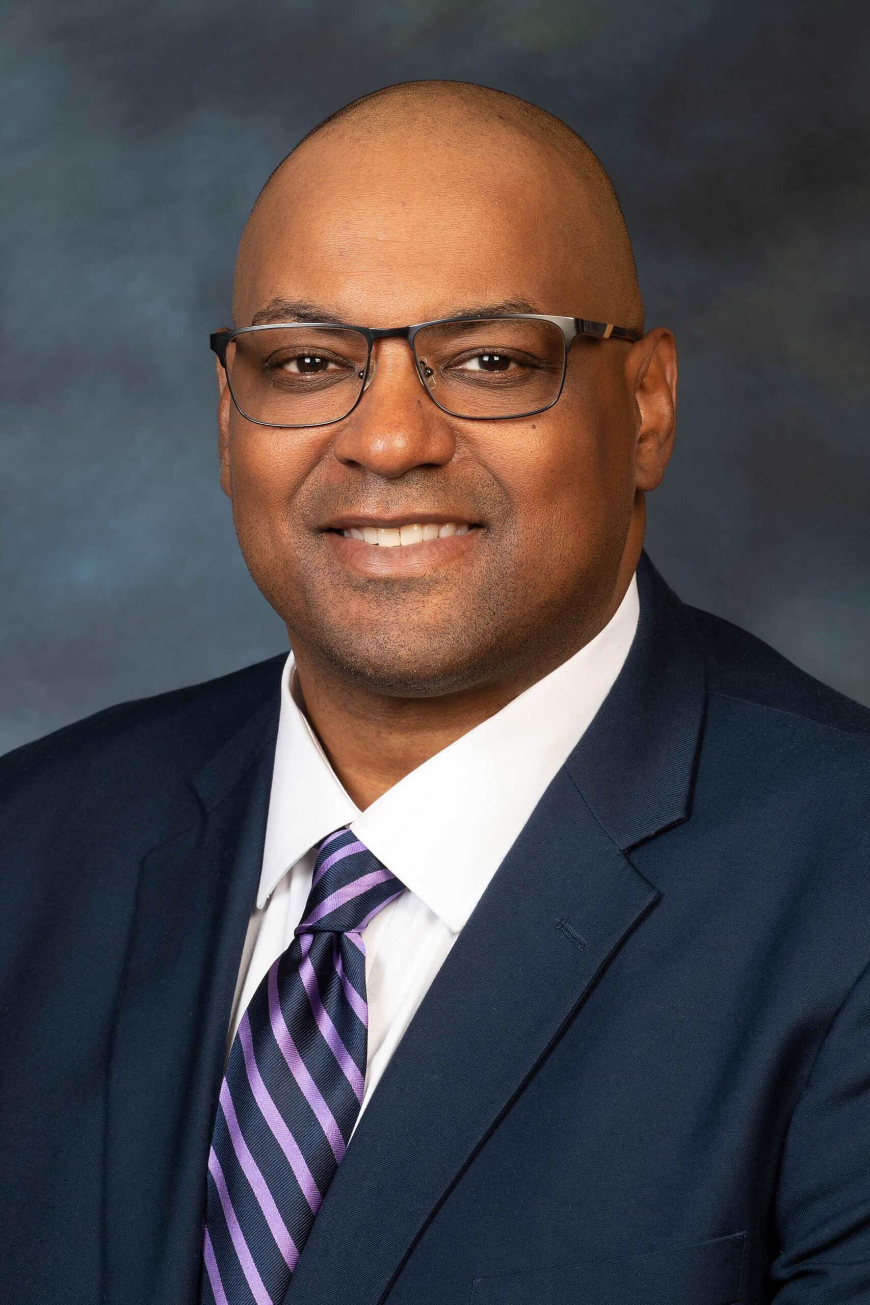 Professional headshot of a city councilmember wearing glasses, a navy suit, white shirt, and purple striped tie, posed against a dark blue gray studio backdrop.