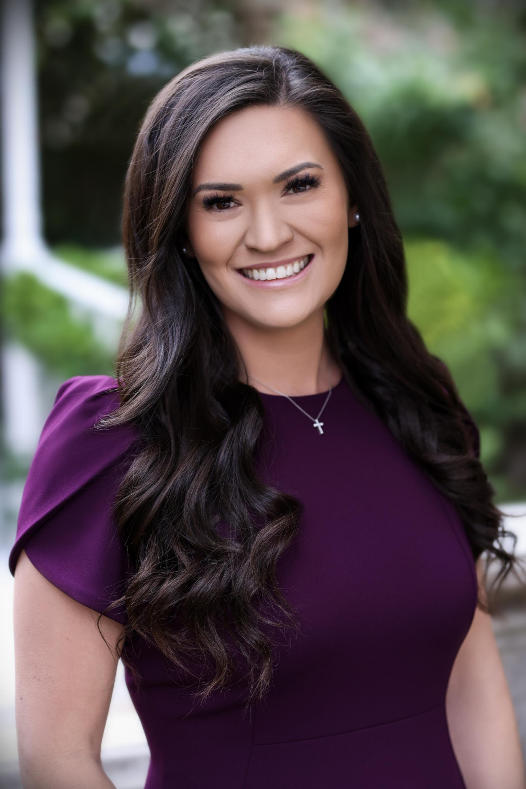 Professional portrait of the vice mayor with long dark hair, wearing a purple dress and cross necklace, smiling outdoors with greenery in the background.