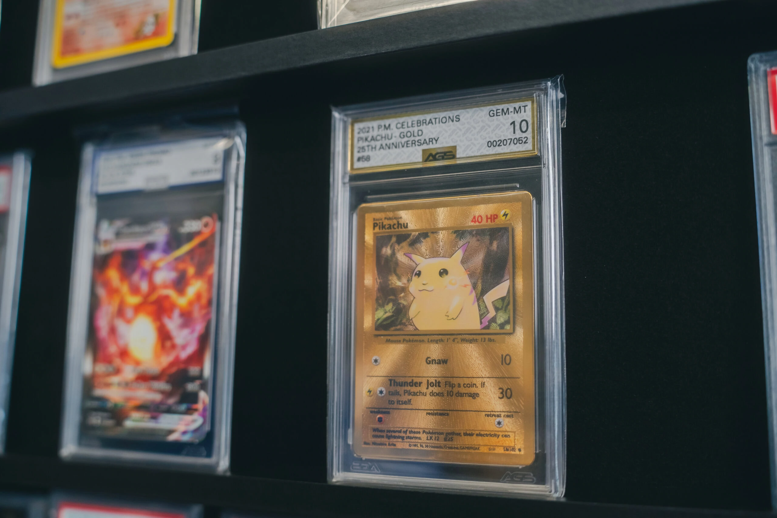 A graded gold Pikachu 25th Anniversary Pokémon card displayed on a black shelf alongside other protected trading cards