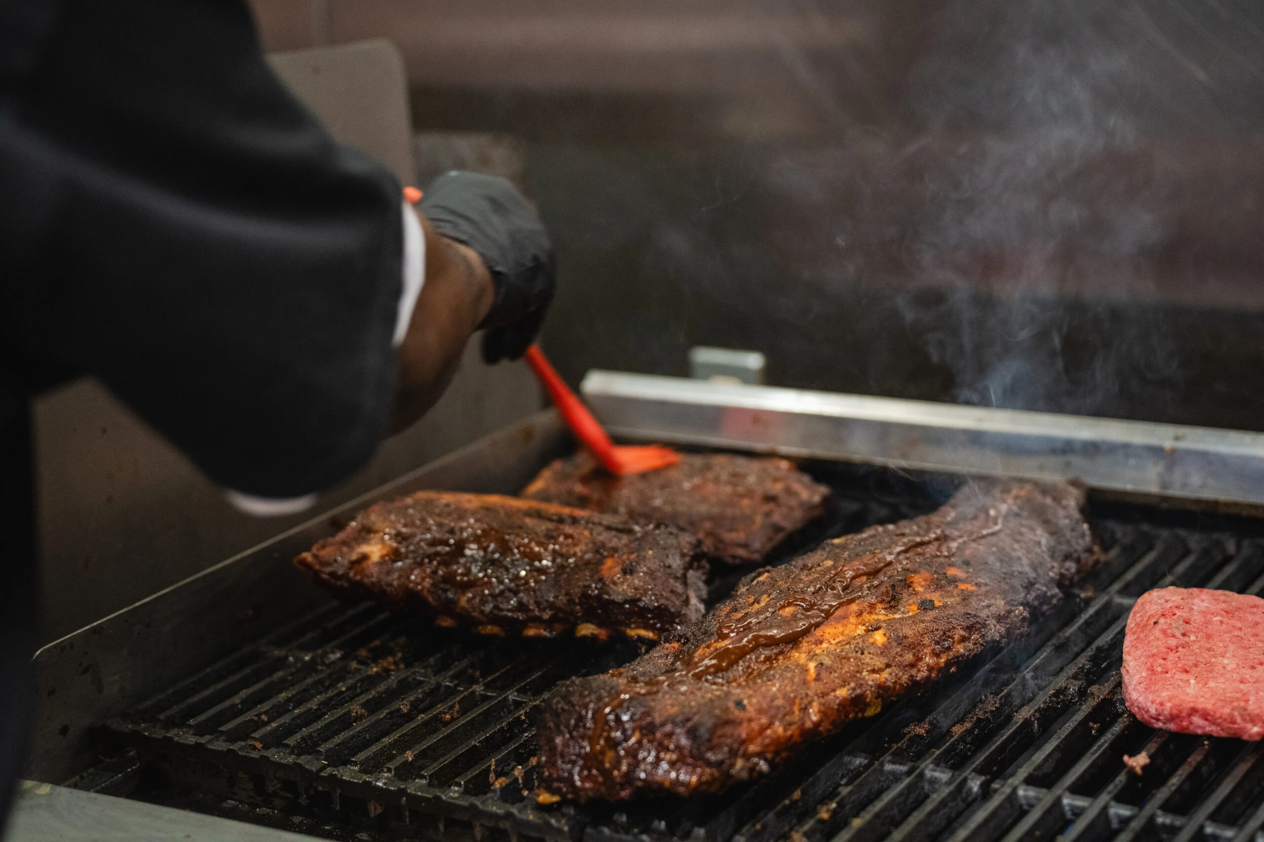 A cook brushes barbecue sauce onto racks of ribs as they smoke on the grill.
