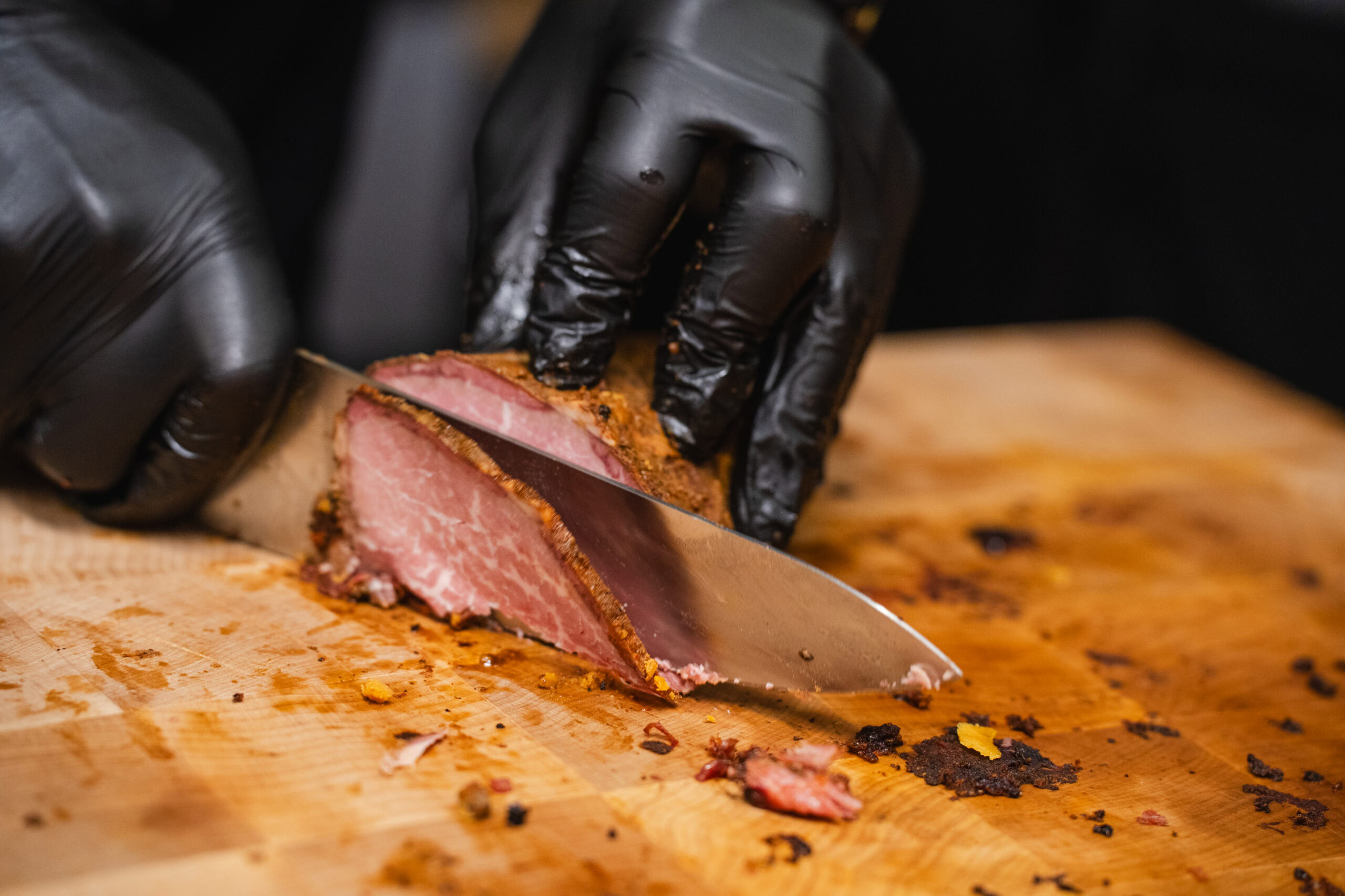 Gloved hands slice smoked meat with a seasoned crust on a wooden cutting board.