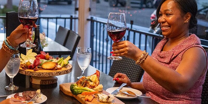 Two women enjoying dinner and raising wine glasses for a toast at an outdoor restaurant table. Various foods, including a charcuterie board, are spread across the table.