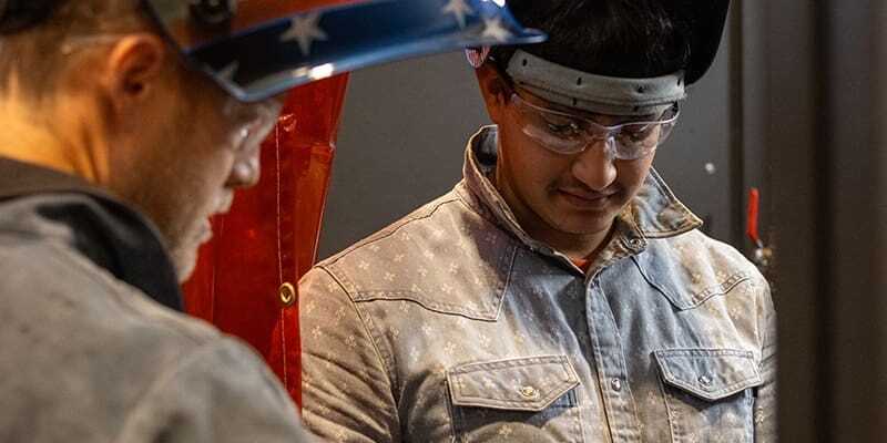 Two people wearing protective welding helmets and safety glasses work on a project together. One adjusts equipment while the other observes.